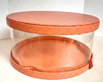 May include: A clear, round display case with a peach-colored, quilted top and base. The sides are transparent, allowing a full view of the contents. The case has a small, peach-colored tab on the side. The case is designed for displaying items.