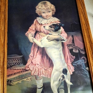 May include: A framed print featuring a child with blonde hair wearing a pink dress with a white ruffled collar, standing and holding a white dog with black spots and a black patch over one eye. The frame is made of light-colored wood.