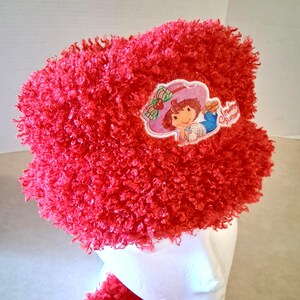 May include: A bright red fuzzy textured hat featuring a circular patch with an illustration of a character wearing a pink hat and blue pants, with the text Strawberry Shortcake printed in red script on the right side of the patch.