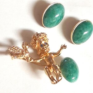May include: A gold-toned brooch featuring a cherub holding a bird, with two green jade oval earrings. The earrings have gold-colored rims. The jade has a glossy finish. The jewelry is set against a white background.