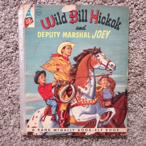 May include: A vintage children's book titled "Wild Bill Hickok and Deputy Marshal Joey." The cover illustration depicts cowboys on horseback. The book is a Rand McNally Book-Elf Book.