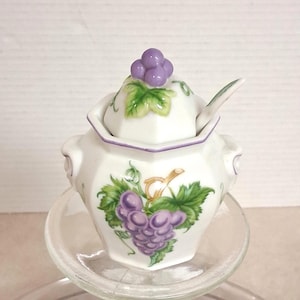 May include: A white ceramic sugar bowl with a lid and spoon, decorated with purple grapes and green leaves. The lid is topped with a cluster of grapes. The bowl sits on a clear glass plate.