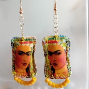 Puede incluir: Pendientes hechos a mano con un retrato de Frida Kahlo. Los colgantes rectangulares de tela están cosidos con hilo amarillo y adornados con una franja de cuentas amarillas. Los pendientes cuelgan de ganchos dorados.