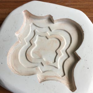 May include: A white, oval-shaped mold with a layered, ornate design. The interior of the mold features a repeating pattern with a light brown hue, while the outer edges are a clean white. The mold appears to be made of a durable material.