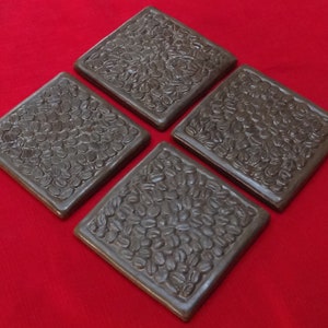 May include: Four square brown ceramic coasters with a raised design of coffee beans.