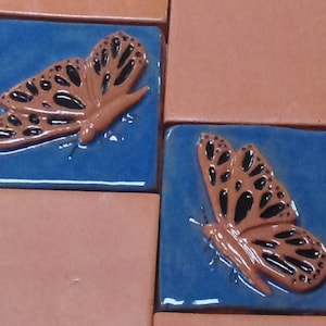 May include: Two ceramic tiles with a blue background and a brown butterfly design. The butterflies have black markings and are in a raised design.