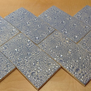 May include: A collection of rectangular ceramic tiles arranged in a herringbone pattern. The tiles are a muted blue-gray color with a textured, bubbly surface. The tiles are approximately 4 inches by 8 inches.
