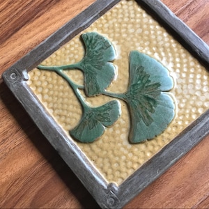 May include: A square ceramic tile featuring a raised relief of three green ginkgo leaves set against a textured yellow background with a honeycomb pattern. The tile is framed by a dark gray border with small circular indentations at the corners.