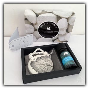 May include: A black gift box containing a white clay diffuser stone with a leaf design, a small bottle of essential oil, and a white tag that says "Just for you". The box is decorated with a white background and white stones. The box has a label that says "Aroma Stones Handmade Clay Diffuser Stones with Fragrance Oil".