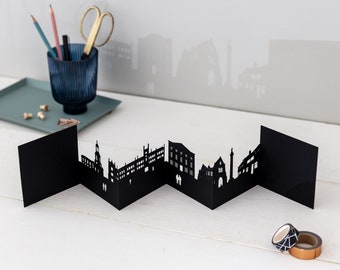 May include: Black paper cityscape model with a zig-zag design. The model is made of multiple panels that fold out to create a 3D effect. The cityscape features buildings and a tower.