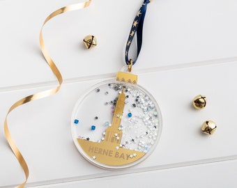 May include: Gold Christmas ornament with a clear plastic sphere filled with silver glitter and blue and gold stars. The ornament features a gold silhouette of a building with the text "HERNE BAY" on the bottom.