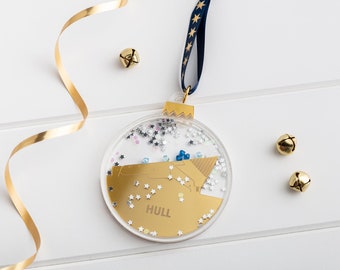 May include: A clear glass Christmas ornament with a gold glitter background and a gold outline of the city of Hull, England. The ornament is decorated with blue and silver glitter and has a blue ribbon attached.
