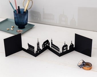 May include: Black paper cityscape silhouette card with a zig-zag fold design. The card features a series of buildings, including a church, a tower, and a few houses.