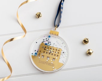 May include: Gold Christmas ornament featuring a building with the word "SHEFFIELD" underneath. The ornament is filled with glitter and stars and has a blue ribbon.