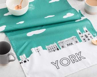 May include: A teal tea towel with a white outline of the York skyline and the word "YORK" in black. The tea towel has a white background with a teal cloud pattern.