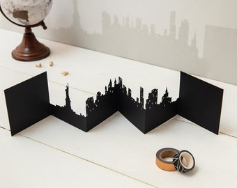 May include: A black paper cityscape card with a silhouette of the New York City skyline, including the Statue of Liberty. The card is folded into sections and is displayed on a white wooden surface.