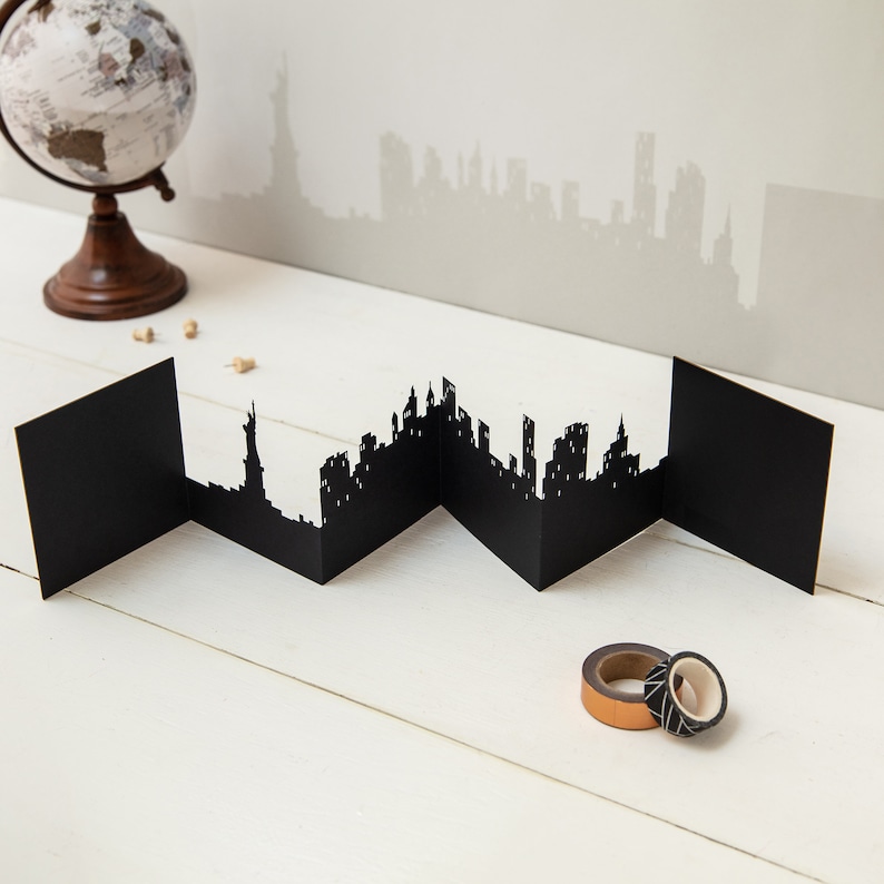 May include: A black paper cityscape card with a silhouette of the New York City skyline, including the Statue of Liberty. The card is folded into sections and is displayed on a white wooden surface.