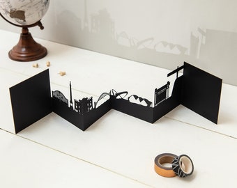 May include: Black paper cut-out cityscape of a city skyline, folded into a zig-zag pattern. The cityscape is on a white wooden surface with two rolls of tape.