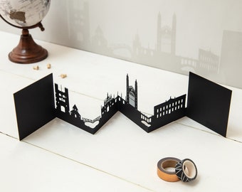 May include: A black paper cut-out of a city skyline, featuring detailed architectural silhouettes. The artwork is displayed on a white surface, with a globe and decorative tape rolls in the background. The skyline is a zig-zag design.