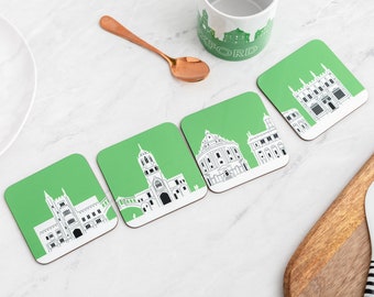 May include: Four green square coasters with white outlines of famous buildings in Oxford, England. The coasters are arranged in a row on a white marble surface. The text "Oxford" is visible on a white mug in the background.