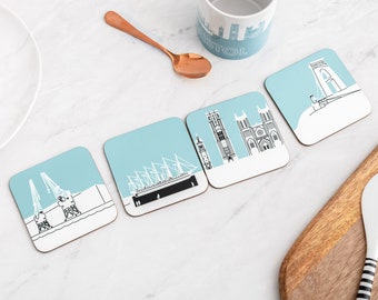 May include: Set of four blue and white coasters featuring different landmarks of Bristol, England. The coasters are made of cork and feature a blue background with white outlines of the landmarks. The landmarks include the Clifton Suspension Bridge, the Bristol Cathedral, the SS Great Britain, and the Bristol Harbourside.