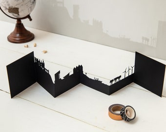 May include: Black paper cityscape silhouette card with cutouts of buildings, wind turbines, and ships. The card is folded into a zig-zag pattern.