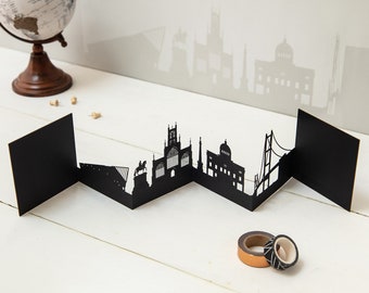 May include: Black paper cityscape model of a city skyline with buildings and a bridge. The model is folded into a zig-zag shape and is sitting on a white wooden surface.