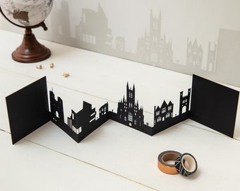 May include: Black paper cut-out cityscape of a city skyline with tall buildings and a church. The cityscape is folded into a zig-zag pattern.