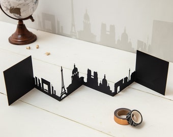 May include: Black paper cityscape silhouette of Paris, France, with the Eiffel Tower, Notre Dame Cathedral, and the Arc de Triomphe. The silhouette is folded into a zig-zag shape and stands on a white surface.