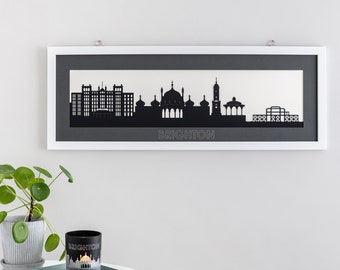 May include: A black and white silhouette of the Brighton skyline, including the Royal Pavilion, the Brighton Pier, and other notable buildings. The silhouette is framed in a white frame with a gray mat. The word "BRIGHTON" is printed in white below the silhouette.
