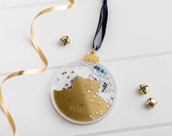 May include: Gold Christmas ornament with a silhouette of York Minster inside. The ornament is filled with silver and blue glitter and has a blue ribbon attached.