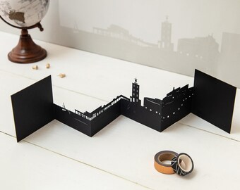May include: Black paper cityscape model with a zig-zag design. The model is made of multiple panels that fold together to create a 3D effect. The model is on a white surface.
