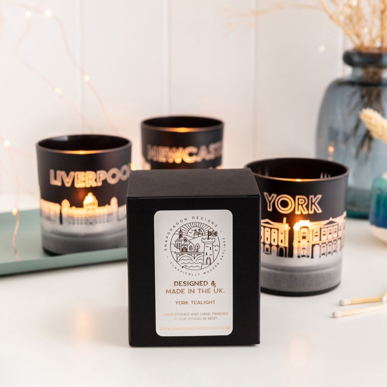 May include: Black glass tealight holders with city skylines of Liverpool, Newcastle, and York. A black box with the text "Designed & Made in the UK" and "York Tealight" is in the foreground. The tealights are lit, and the background is blurred.