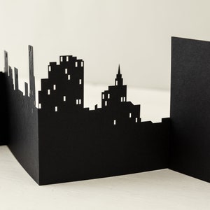 May include: Black paper cityscape silhouette with buildings of varying heights, including a tall skyscraper and a church steeple. The cityscape is cut out of a single sheet of paper and stands on a white surface.