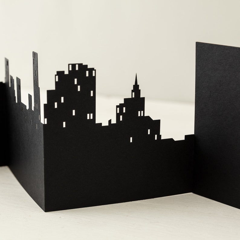 May include: Black paper cityscape silhouette with buildings of varying heights, including a tall skyscraper and a church steeple. The cityscape is cut out of a single sheet of paper and stands on a white surface.