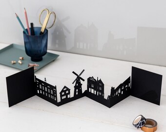 May include: Black paper cut-out cityscape card with a windmill, houses, and buildings. The card is folded and stands upright on a white surface.