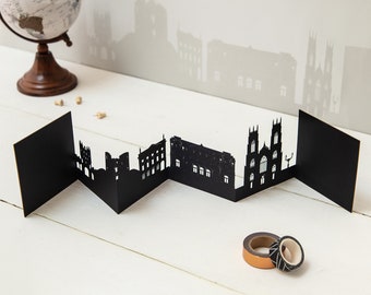 May include: A black paper cityscape silhouette with a zig-zag design. The paper art features building outlines and is displayed on a white surface. A small globe and decorative tape rolls are also present.