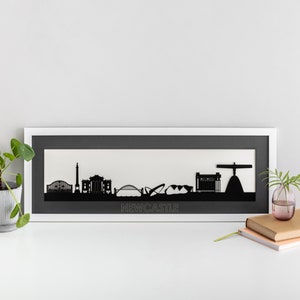 Puede incluir: Una silueta recortada en papel negro y blanco del horizonte de Newcastle, enmarcada en un marco blanco con un paspartú gris. El horizonte incluye el puente Tyne, el Sage Gateshead y el Ángel del Norte. La palabra "NEWCASTLE" está impresa en gris debajo del horizonte.