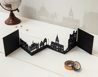 May include: A black paper cityscape card with a folded design. The card features a silhouette of a city skyline with buildings and a church.