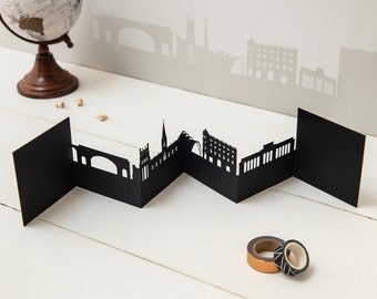 May include: Black paper cityscape card with a folded design. The card features a silhouette of a city skyline with buildings and bridges.