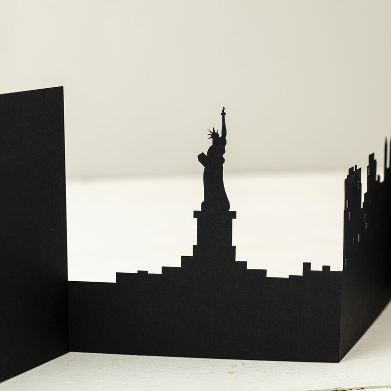 May include: Black paper cutout of the New York City skyline, featuring the Statue of Liberty and skyscrapers.