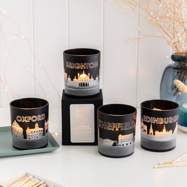 May include: Four black glass candle holders with city skylines: Oxford, Brighton, Sheffield, and Edinburgh. Each has a gold-colored city name and skyline design. The candles are lit, creating a warm glow. The set includes a black box.