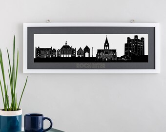 May include: Black and white silhouette print of the Rochester skyline, featuring buildings and a castle. The print is framed in a white frame with a gray mat. The word "ROCHESTER" is printed below the skyline.