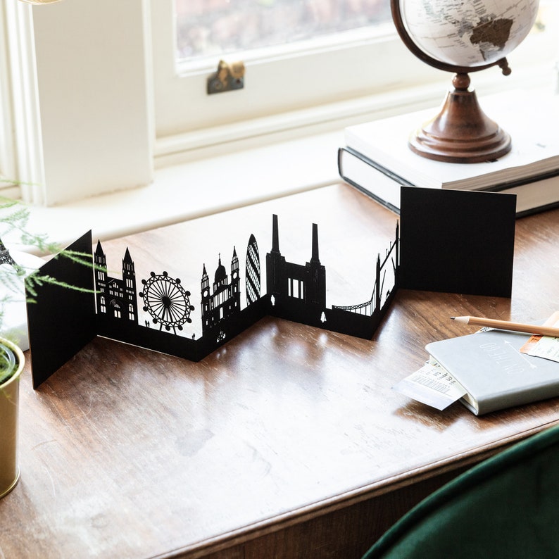 May include: A black papercraft cityscape of London, England, with iconic landmarks like the London Eye, Big Ben, and the Houses of Parliament. The cityscape is folded into a series of panels, creating a three-dimensional effect.