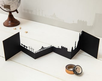 May include: A black paper card with a cut-out silhouette of a city skyline. The card is folded in a zig-zag pattern, creating a 3D effect. Two rolls of washi tape are in the foreground.