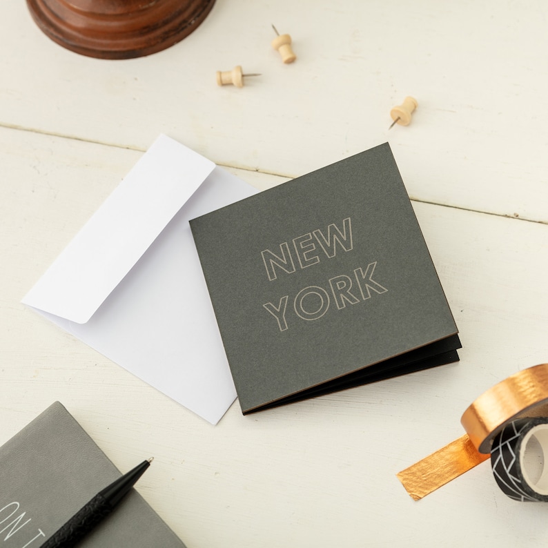 May include: A dark gray card with the words "NEW YORK" in white outlined letters. The card is folded in half and is sitting on a white surface. There is a white envelope and a few push pins on the surface.