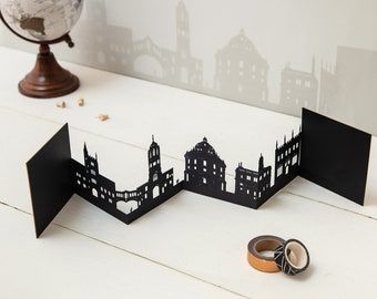 May include: Black paper cut-out cityscape card with a folded design. The card features a silhouette of a city skyline with buildings and a bridge.