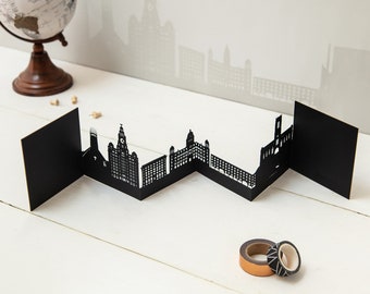 May include: Black paper cut-out cityscape of Liverpool, England. The cityscape is folded into a zig-zag pattern and is displayed on a white surface.