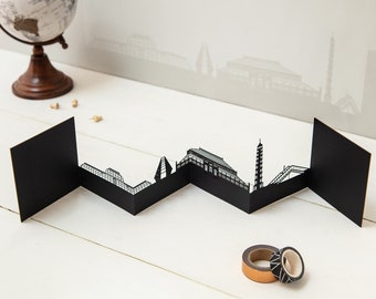 May include: A black paper cityscape card with a cut-out design of a city skyline. The card is folded into a zig-zag shape and is sitting on a white surface.