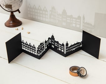 May include: Black paper cityscape model with a zig-zag design. The model is made of multiple panels that fold together to create a 3D effect. The cityscape features buildings with different heights and shapes.